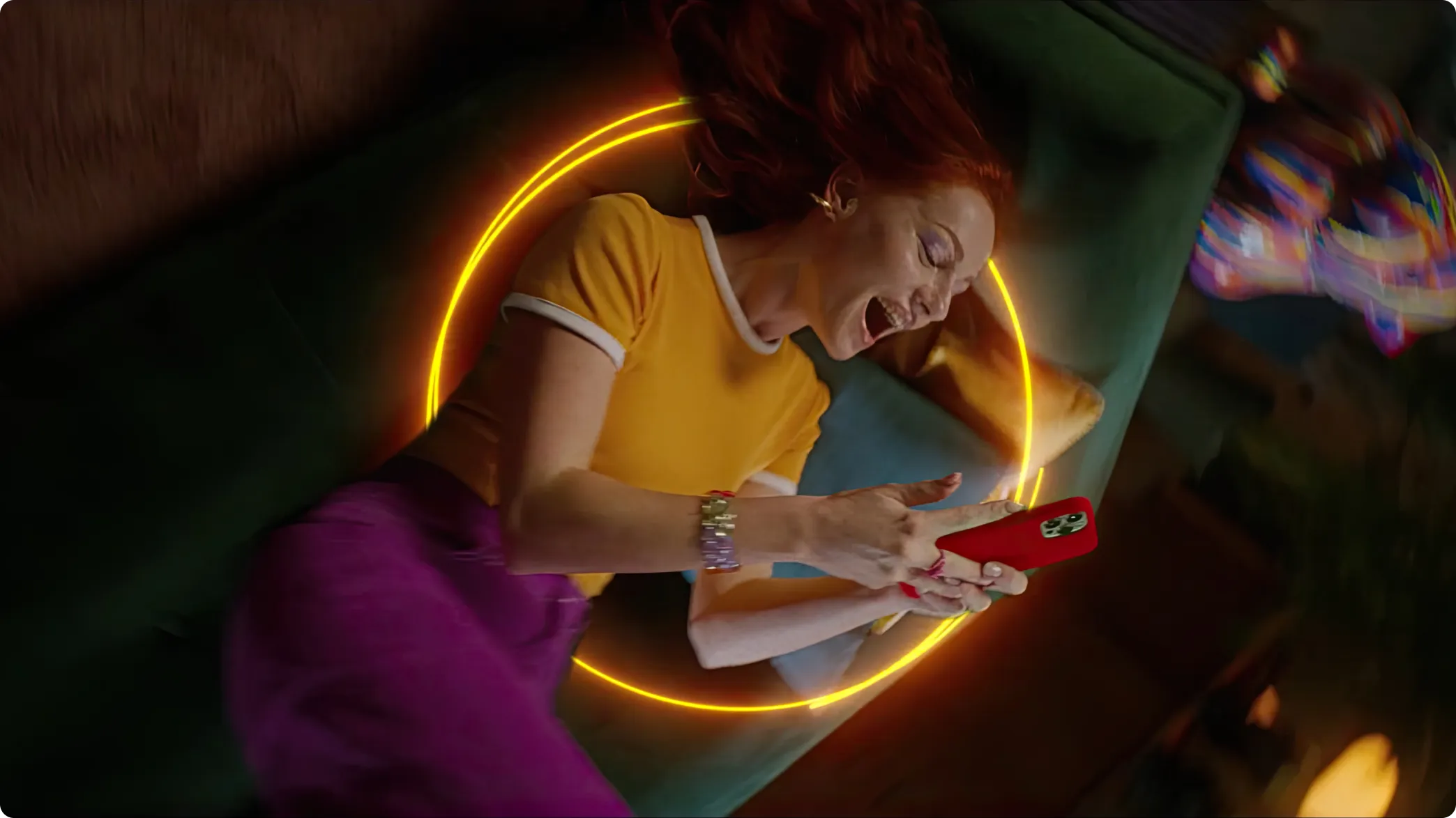 Young red-haired woman laughing on a green couch, looking at her smartphone. Wearing a yellow t-shirt and purple pants, surrounded by a glowing yellow circle and colorful, blurred lights.