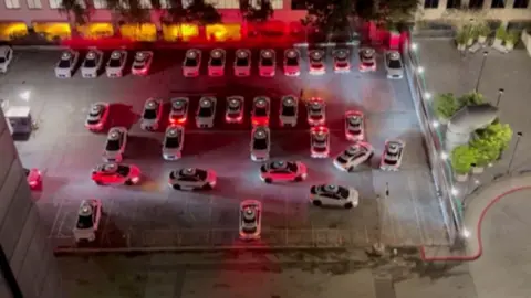 Rows of cars line a car park at night with red lights on and some in a queue