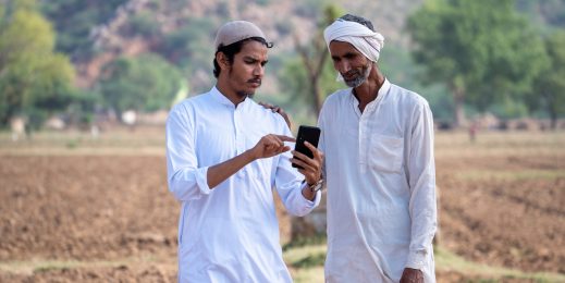 Two farmers standing in an arid village field looking at a mobile phone.