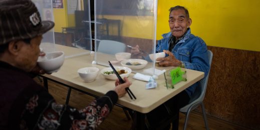 Two elderly men sit across a table, each holding a bowl and chopsticks 