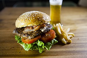 A brown meat patty between slices of yellow American cheese, topped with sliced pickles and lettuce, sandwiched in a sesame seed bun