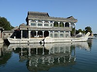 City lake, about 200 m in diameter, with a fountain in the center that shoots water about 25 m high. A few small pleasure craft are in the lake. In the background are trees and buildings.