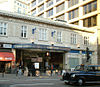 A light grey building with "M.R. ALDGATE STATION M.R." written in stone on the front face and a black car driving in the foreground