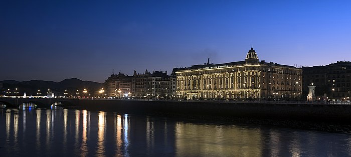 Vista del Hotel María Cristina, desde la orilla del río Urumea en el anochecer. Cielo azulado y reflejos de luces cálidas (amarillas) y blancas en el río.