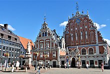 Frontale Farbfotografie von zwei weiß-braunen Gebäuden mit verzierten Staffelgiebeln. An jeder Staffelecke stehen Figuren und kleine Obelisken. An den Dachspitzen befinden sich Eisenkreuze. An der rechten Fassade hängen vier Statuen und vor dem rechten Gebäude ist ein flacher Vorbau mit einem verzierten Torbogen, in dessen Pfeilern zwei Farbfiguren stehen. Auf dem Vorplatz befindet sich eine weiße Ritterstatue mit Wappenschild.