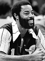 A man, wearing red and green jacket with a red shirt and tie, is standing on a basketball court while holding a microphone.