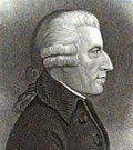 A profile view of a pleasant faced clean shaven man in his 50s or 60s with a strong nose. He is wearing a gray colored curly wig with a pony tail, and a tweed jacket with large lapels and buttons.