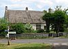 Thatched stone house surrounded by trees. In the foreground a road junction and sign.