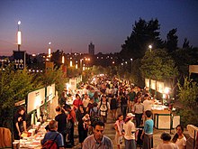 An outdoor book fair at dusk. Vendors are in booths along both sides of a walkway crowded with people.