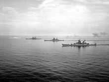 A black and white photograph of four large ships sailing on calm seas from the right to the left.