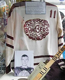 White hockey jersey with maroon trim and a Bombers logo on the chest, accompanied by a hockey stick, a hockey glove ,and a photo of Bobby Clarke