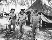 Three men wearing military uniforms walking towards the camera. A tent and palm trees are in the background