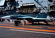 Aqua and blue jet aircraft on aircraft carrier deck, with a group of men standing close-by. Behind the jet is the ship's island