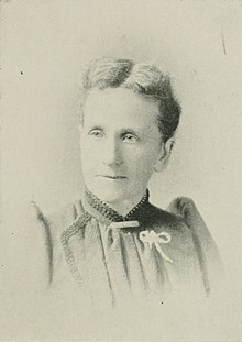 B&W portrait photograph of a middle-aged woman with hair in an up-do wearing a high-collared blouse with a white ribbon pinned on the left side.