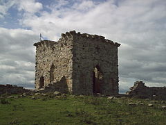 A tall stone built ruined square building. At the top are visible parts of the battlements. In the foreground a grassy slope with scattered stones leading up to the building. In the background white clouds with a small patch of blue sky.