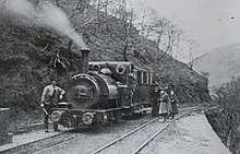 A train consisting of a locomotive and single carriage stands in the centre track of three tracks, which is on a ledge on a steep hillside. There is no platform. Two adults and two girls in Victorian dress stand on the track beside the carriage, with a man leaning against the locomotive and another man on the footplate.