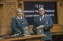 Un homme avec des lunettes et une femme blondes, vêtus d'un uniforme vert-gris, tenant chacun une boîte métallique estampillées d'une croix blanche sur fond rouge, se tenant devant un panneau synoptique.