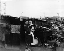 Photographie en noir et blanc d'une femme avec un habit traditionnel japonais portant un petit enfant devant une baraque hâtivement construite. Des tas de décombres et des maisons intactes sont visibles à l'arrière-plan.