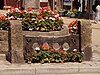 Stocks, made from central dark wooden stock beams with two holes in. The beams are supported by stone pillars. They have flowers planted in front of them, and the market cross, also with flowers, sits behind.