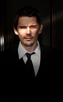 A Caucasian male with dark brown hair and stubble, wearing a black suit with a white shirt and black tie.