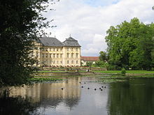 Frontale Farbfotografie eines dreigeschossigen, hellgelben Schlosses mit grauem Dach. Die linke Seite ist von einem Baum verdeckt. Im Vordergrund sind Terrassen mit Blumenbeeten und eine Wiese. Die Wiese ist mit einer Steinmauer vom Wasser getrennt, auf der Enten und Seerosen schwimmen. Rechts hinten sind eine Nebengebäude und Bäume.