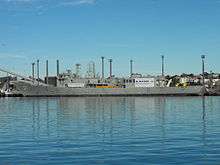 Adelaide tied up alongside at a wharf. The majority of her communications and radar masts have been removed, she carries no weapons, and several large squares have been cut into the ship's hull.