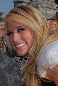 A close-up photo of a young, Caucasian blonde, who is smiling at the camera. Men in camouflage uniforms are visible in the background.