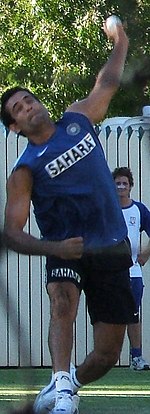 Young brown skinned clean shaven man wearing a blue sleeveless T-shirt with the word "SAHARA" in black, wearing dark shorts is about to deliver a white cricket ball. His left bowling arm is above his head and he is wearing white sports shoes. His leading right leg is straight while his trailing left leg is bent. He is grimacing. In the background is a white picket fence and shady green vegetation.