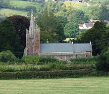 Gray stone building with square tower surmounted by a spire on the left. Surrounded by trees and green fields.