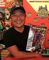 An Asian man in a black hat and T-shirt poses with a comic book.