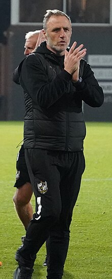 A white middle-aged male in a Port Vale F.C. tracksuit clapping his hands to applaud the crowd at Vale Park