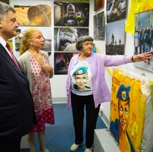 Ein Mann und zwei Frauen stehen auf der linken Bildseite in einem Raum mit vielen Fotos und Objekten an den Wänden. Eine Frau (Savchenko) zeigt mit ausgestrecktem Arm auf ein Bild.