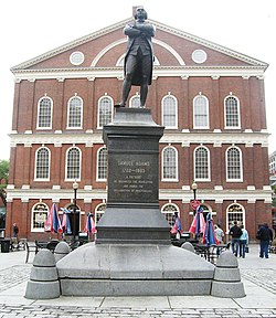 A statue on a pedestal of a man standing with his arms crossed. An inscription on the pedestal reads, "Samuel Adams, 1722–1803. A Patriot. He organized the Revolution and signed the Declaration of Independence." Behind the statue is a three story brick building with many windows.