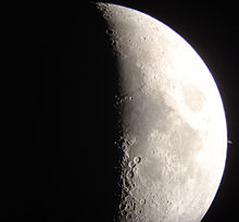 A Lua crescente, iluminada no seu lado direito, ocupa o centro da imagem. Próximo a ela, no lado direito, Saturno, muito menor, coloração amarelada.