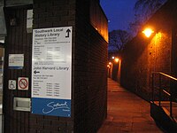 Evening scene looking into an alleyway, bounded on the right by a long, 15-foot-high brick wall with brightly-lit lamps hanging on it. On the left, there is a building with a sign saying "John Harvard Library", with telephone numbers and opening hours. An arrow points toward the entrance to the library in the alleyway.