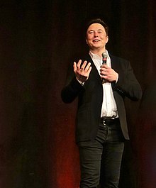 Musk speaking in front of curtains; he is holding a microphone.