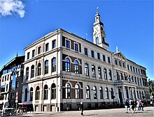 Farbige Eckansicht eines dreigeschossigen Gebäudes mit einem Säuleneingang, über dem sich ein Balkon mit Fahnen befindet. Über dem Giebelfeld steht auf dem Dach eine Figur. In der Dachmitte ist eine Turmhaube mit Uhr und die meisten Fenster haben Rundbögen.