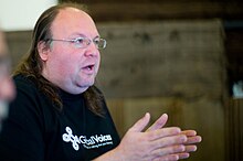 A photograph of a double chin spectacled man with hair slicked back speaks