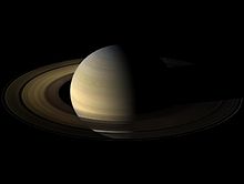 Saturno no centro da imagem, com sua metade esquerda iluminada, com coloração bege. Os anéis aparecem com formato elíptico, devido à visão obliqua, marrons.