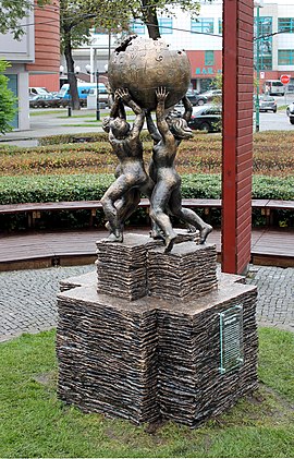 Four figures stand in a circle holding up the symbol of Wikipedia, which consists of a globe made of jigsaw pieces, with few pieces missing, symbolizing that the work of the encyclopedia will never be finished