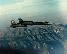 Sideview of black jet aircraft overflying mountain towards right of photo.