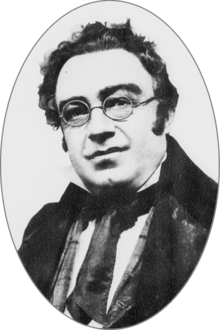 Round-faced white man, clean shaven with unruly dark hair and round spectacles