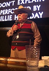 Un homme avec une grande moustache grise, un chapeau de cowboy et un pull rayé horizontalement rouge et noir. Il tient un micro dans sa main droite.