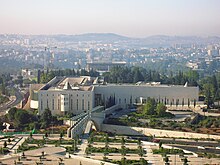 A low-rise T-shaped building with small, slit-like windows, with a small park in front, and a hilly, inhabited terrain in the background