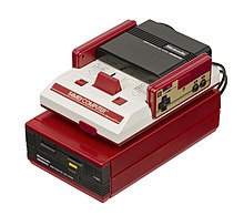 Un'unità Famicom bianca e rossa si trova in cima a un'unità Famicom Disk System rosso caramella con unità disco inseribile nera. Due controller rettangolari, ciascuno con un D-pad e due pulsanti neri, si inseriscono nel Famicom.