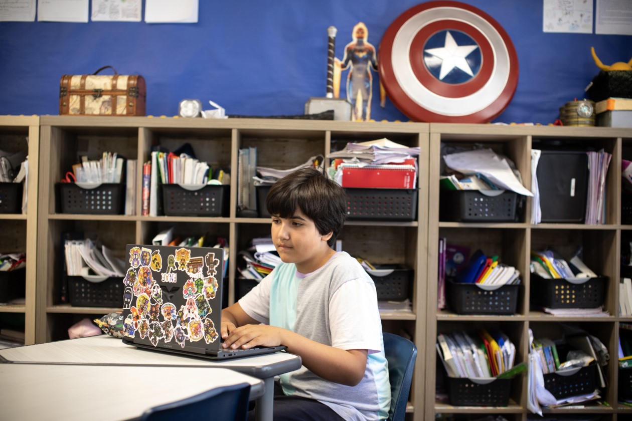 Decorative. A student sitting at a desk in a classroom and working on a laptop. There are bookshelves with bins of notebooks and folders behind the student.